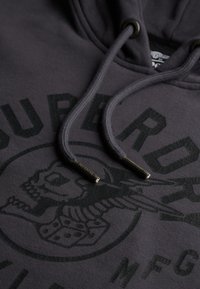 Donkergrijze hoodie met een gestructureerd zwart grafisch ontwerp van een schedel en vleugels, afgewerkt met bijpassende koorden en gelabelde metalen punten.