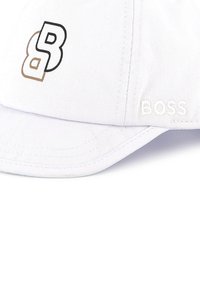 White cap made of cotton with a structured shape. Features black and gold embroidered logo and brand name on the front left side.