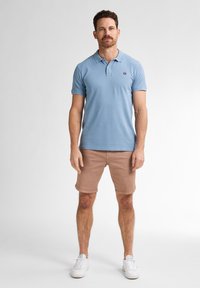 Lichtblauwe polo met een kraag en korte mouwen, gecombineerd met lichtbruine shorts en witte sneakers. Eenvoudig logo detail.