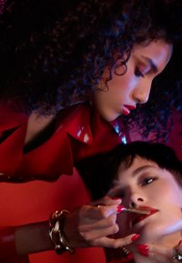 Deux individus aux cheveux noirs et bouclés et aux cheveux courts, portant un outfit rouge brillant, appliquent du rouge à lèvres rouge. Ils sont devant un fond rouge vif.