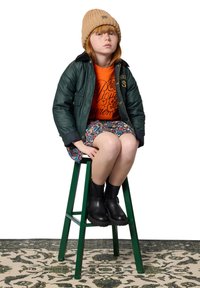 Veste matelassée verte avec col noir, sweat-shirt graphique orange, jupe à motifs floraux, bonnet en tricot beige et bottines noires. Assise sur un tabouret vert.