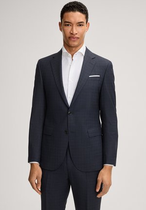 Mężczyzna w ciemnoszarym garniturze na dwa guziki z dopasowanymi spodniami i białą koszulą, stoi na tle jasnego, jednolitego tła.