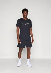 Nike urheiluasu, jossa on tummansininen pyöreäkauluksinen t-paita hopeisella logolla ja yhteensopivat shortsit. Materiaali vaikuttaa kevyeltä ja sileäpintaiselta.