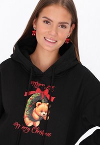 Fekete kapucnis pulóver, amelyen koszorúban lévő mackó grafikája látható, piros részletekkel és a "Boldog Karácsonyt" szöveggel játékos betűtípussal. Piros masnis fülbevalók.