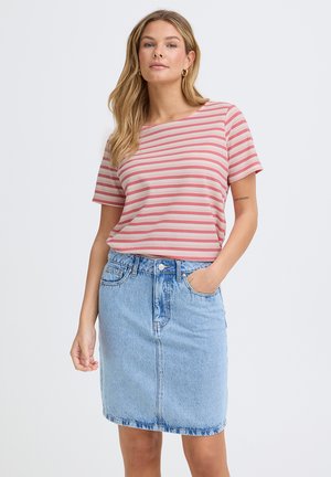 Nő áll, kezét zsebében tartva, rózsaszín csíkos rövid ujjú felsőt és világoskék farmer szoknyát visel egyszerű háttér előtt.