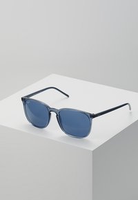 Ochelari de soare pătrați cu nuanță albastră, având un cadru transparent de culoare albastră. Dispun de brațe elegante cu detalii ale logo-ului și margini rotunjite pentru un design minimalist.