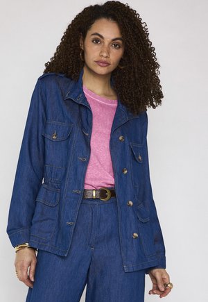 Donna con capelli ricci che indossa una giacca e pantaloni di jeans blu sopra una maglietta rosa, accessoriata con orecchini a cerchio dorati e braccialetti.