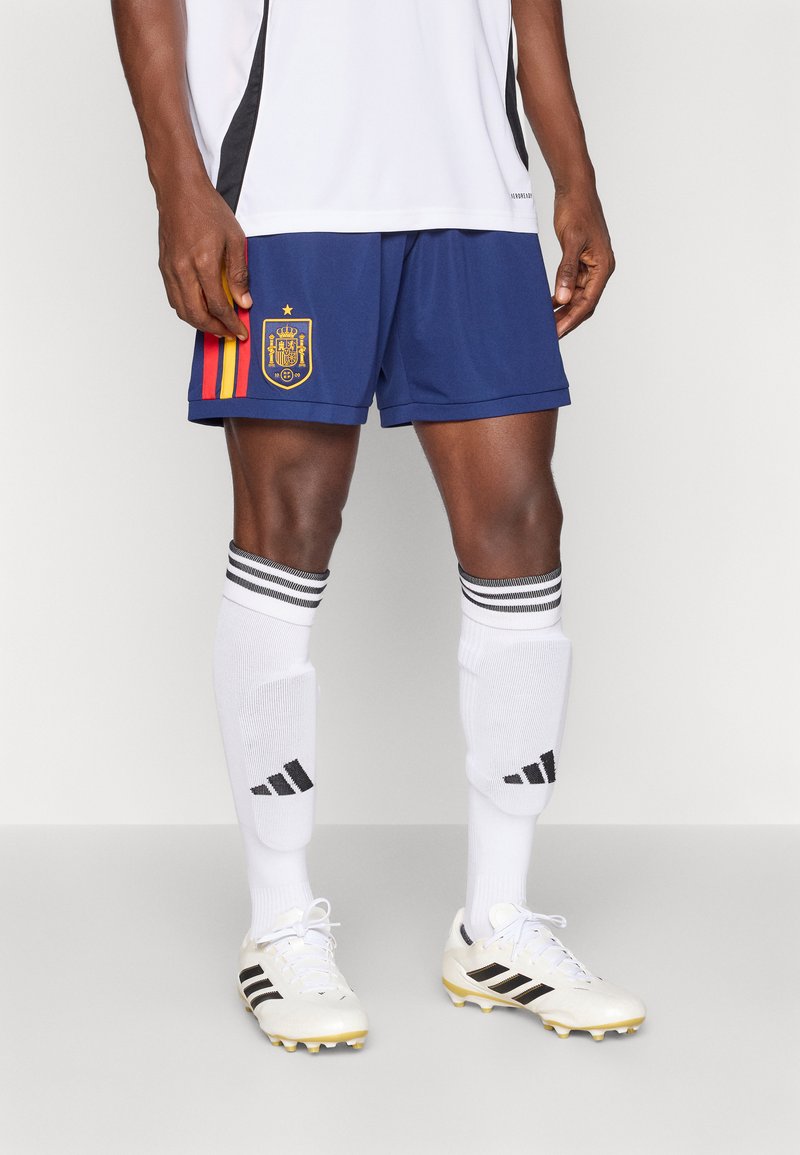 Μπλε ποδοσφαιρικές βερμούδες με κόκκινες και κίτρινες ρίγες, που διαθέτουν σήμα. Συνδυάζονται με λευκές κάλτσες διακοσμημένες με μαύρες ρίγες και λευκά παπούτσια.