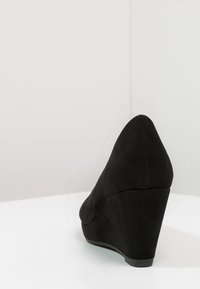 Černá semišová wedge obuv s špičatou špičkou a hladkým povrchem, která se vyznačuje mírným wedge podpatkem a elegantním, štíhlým designem.
