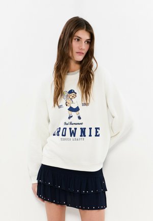 Joven con cabello largo y castaño que lleva una sudadera blanca con un oso jugando al tenis y una falda negra de volantes con tachuelas.