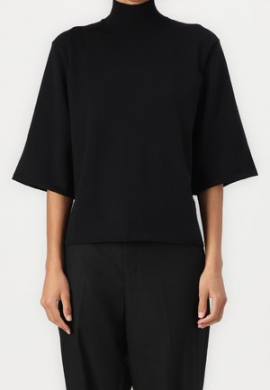 Person iført en sort, højhalset top med brede ærmer til albuen og sorte bukser mod en ensfarvet lys baggrund.