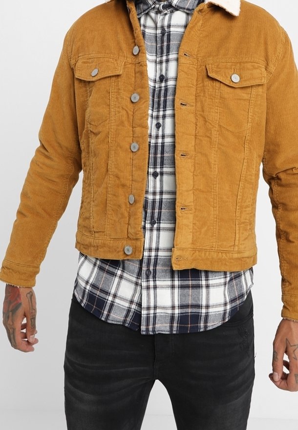 Homme portant une veste en velours côtelé moutarde avec col en sherpa blanc, par-dessus une chemise à carreaux noir et blanc et un jean noir, debout avec les mains légèrement levées.