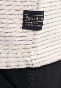 Camisa de punto a rayas en crema y azul marino, con un tejido texturizado y una etiqueta de la marca en azul con letras doradas cosida en el dobladillo.