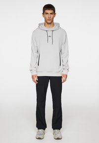 Lys grå hættetrøje med snoretræk i hætten og et logo på brystet, parret med sorte bukser og grå sneakers. Glat stof, afslappet pasform.
