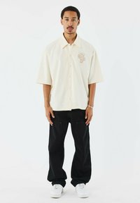 Camisa de botones de manga corta color beige claro con un diseño texturizado en el pecho, combinada con pantalones cargo negros de corte holgado y zapatillas blancas.
