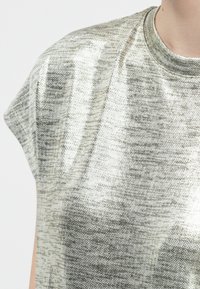 Κοντινό πλάνο ενός ατόμου που φορά μεταλλικό ασημί κοντομάνικο μπλουζάκι με στρογγυλή λαιμόκοψη σε ένα απλό φόντο.