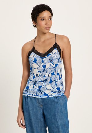 Woman wearing a blue and white floral camisole with black lace trim and loose-fitting blue jeans, standing with one hand in pocket.