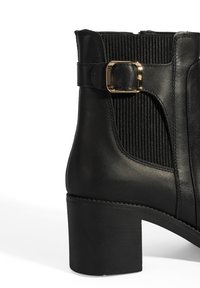 Schwarze Lederstiefelette mit einem dicken Absatz, ausgestattet mit gerippten elastischen Einsätzen und einem goldenen Schnallenakzent an der Seite. Glatte Textur.