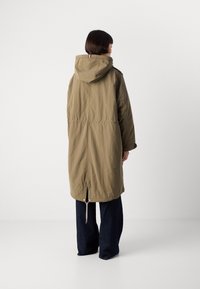 Giacca lunga color khaki con cappuccio, stretta in vita. Presenta un cordoncino sull'orlo e dettagli con bottoni sui polsini. Indossata sopra jeans scuri.