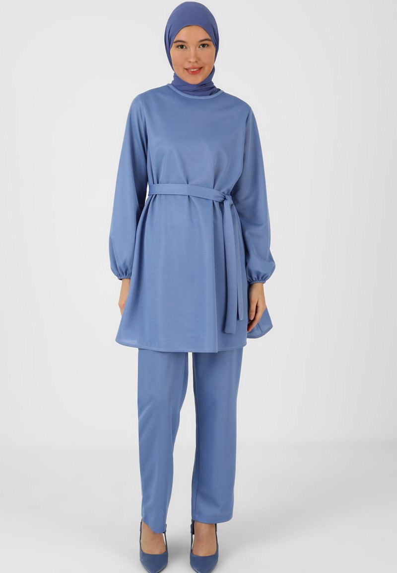 Blaues Zweiteiler-Outfit bestehend aus einer Langarm-Tunika mit hohem Ausschnitt und einem gebundenen Gürtel, kombiniert mit geraden Hosen. Glatte Stoffoberfläche.