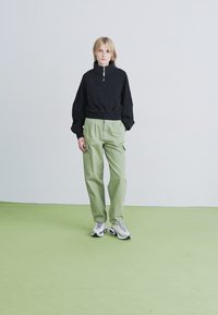 Blonde Frau steht auf grünem Boden, trägt einen schwarzen Pullover mit Reißverschluss, hellgrüne Hose und weiße Sneaker und schaut nach vorne.