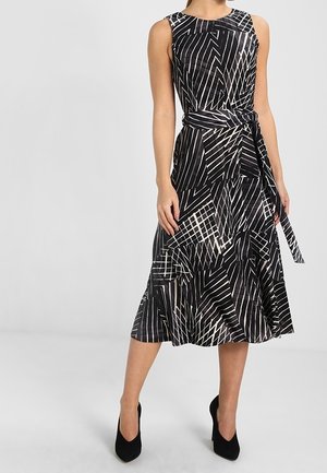 Ärmelloses schwarzes Midikleid mit weißen geometrischen Linien, tailliert mit Gürtel, kombiniert mit schwarzen spitzen High Heels, getragen von einer stehenden Person.