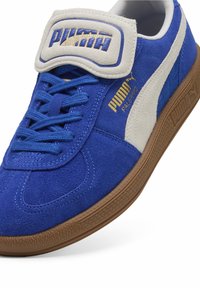 Sneakers PUMA z niebieską zamszową cholewką, białymi paskami akcentującymi, brązową gumową podeszwą oraz rzepem na języku z detalem logo.