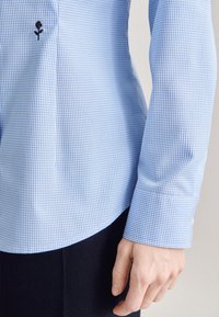 Camicia a scacchi blu chiaro con bottone, dotata di orlo curvo, dettaglio ricamato di rosa nera sul lato sinistro del petto e polsini strutturati con dettagli di bottoni.