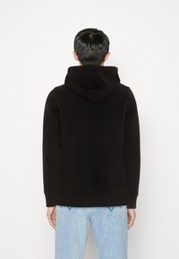 Felpa nera con cappuccio, realizzata in tessuto morbido, presenta una vestibilità rilassata e polsini a coste; indossata con jeans azzurri chiari, che mostrano accenti colorati sulle tasche posteriori.