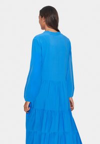 Rochie lungă, de culoare albastră strălucitoare, confecționată dintr-un material ușor. Prezintă mâneci lungi, straturi pliate și un decolteu rotund. Textură netedă cu un luciu subtil.