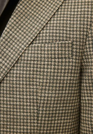 Lähikuva ruskeasta ja beigestä koiranhampaikuviollisesta kankaasta pukutakin kauluksessa, jossa on rintatasku.