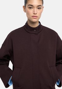Ciemnobrązowa, oversize'owa kurtka z miękkiego materiału, z wysokim kołnierzem, zapięciem na zatrzaski z przodu i bocznymi kieszeniami. Jasnoniebieska koszula pod spodem.