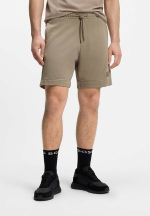 Man met taupekleurige shorts met trekkoord, zwarte sokken met wit logo en zwarte sneakers die op een witte achtergrond staan.