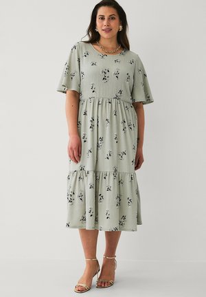 Kvinde iført en lys salviegrøn midi-kjole med sort-hvide blomsterprints, flagrende ærmer og guld stropper på hæle, stående mod en ensfarvet væg.