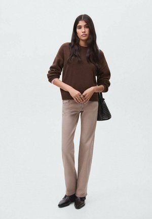 Femme portant un pull brun, un pantalon beige, des bottines noires, tenant un sac à main noir, se tenant devant un fond clair.