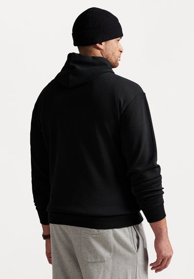 Sweatshirt preto com capuz e acabamento texturizado, corte reto, com mangas longas e um ajuste descontraído. Usado com calças de moletom cinzentas.