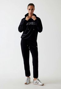 Mustast veluurist dressipluus, millel on kapuutsiga pusa, millel on "GUESS" rhinestone-dega ja sobivad püksid. Välimuse täiendavad hõbedased spordijalatsid.