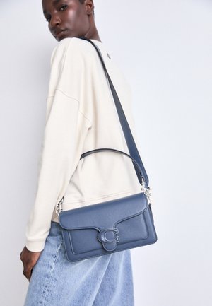 Person iført en cremefarvet sweatshirt og lyseblå jeans, der bærer en blå lædertaske med en stor "C" lukning.