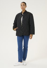Zwarte gewatteerde jas met hoge kraag, drukknoopsluitingen en zijzakken, gecombineerd met blauwe jeans en witte sneakers. Eenvormige achtergrond.