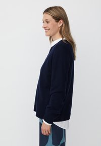 Granatowy sweter z dzianiny z okrągłym dekoltem i prążkowanymi mankietami, nałożony na białą koszulę, noszony z wzorzystymi niebieskimi spodniami.