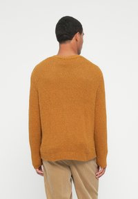 Άνδρας που κοιτάζει μακρι�ά, φορώντας ένα υφασμάτινο μουσταρδί πουλόβερ και μπεζ τζιν καμπάνα, μπροστά σε ένα απλό λευκό φόντο.