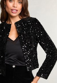 Schwarze Paillettenjacke mit strukturiertem Oberflächen, rundem Ausschnitt und langen Ärmeln, getragen über einem schwarzen Satinoberteil.