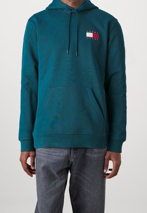 Bluza z kapturem w kolorze morskim, z przednią kieszenią i sznurkami, z małym logo Tommy Jeans na piersi, noszona z wyblakłymi szarymi jeansami.