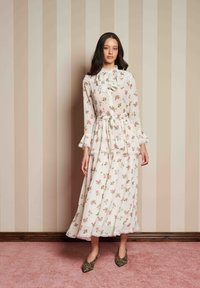 Γυναίκα φορεμένη με λευκό floral φόρεμα μακρυμάνικο με φουσκώματα και ζώνη, στέκεται σε ροζ χαλί μπροστά από καφέ ριγέ τοίχο, με παπούτσια λεοπάρ.