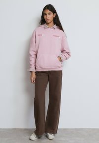Sudadera rosa con capucha, con un bolsillo delantero y puños a rayas, combinada con pantalones marrones de pierna ancha y zapatos de color claro.
