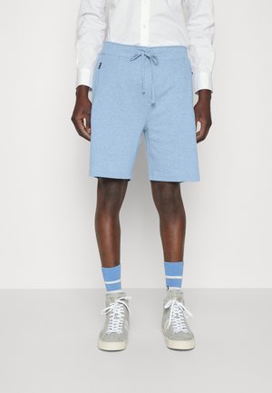 Homme portant un short bleu clair à cordon, une chemise blanche boutonnée, des chaussettes à rayures bleues et des baskets montantes grises, debout sur un fond uni.