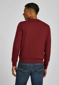 Bordeaux sweatshirt met lange mouwen, ribgebreide manchetten en onderrand, zachte textuur, gecombineerd met blauwe jeans met gele stiksels op de achterzakken.