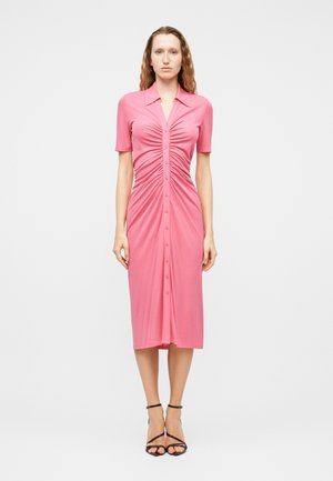 Mujer vistiendo un vestido midi rosa con fruncidos y botones, de manga corta, y sandalias negras de tacón con tiras, de pie frente a un fondo liso.