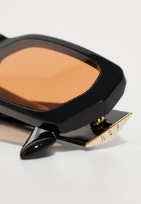 Gafas de sol negras con montura sobredimensionada y angular. Las lentes son de un tono naranja cálido, con herrajes dorados marcados con "RSF".