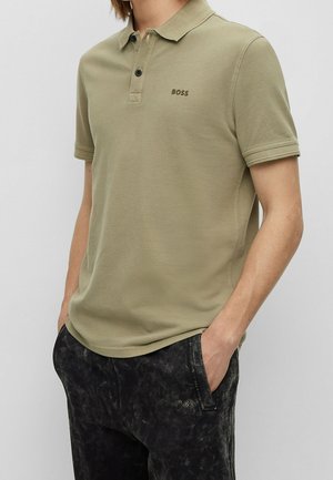 Mężczyzna ubrany w oliwkową koszulę polo z krótkim rękawem z logo "BOSS" oraz czarne, teksturowane spodnie, z rękami w kieszeniach, na jednolitym tle.